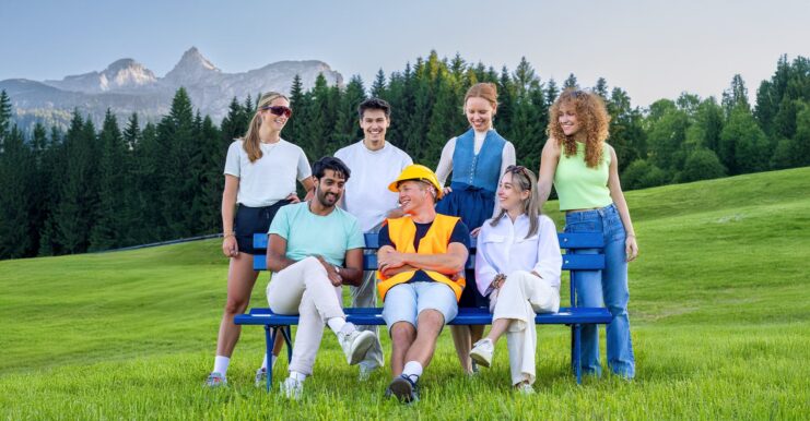 Sieben Jugendliche sind auf einem grünen Feld. Im Hintergrund Berge und ein Wald. Drei sitzen auf einer Bank, vier stehen dahinter. Sie wirken glücklich.