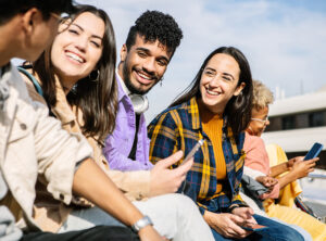 Gruppe junger Menschen sitzt draußen in der Sonne, lacht und schaut auf Smartphones