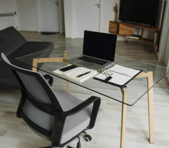 Modernes Homeoffice mit Glastisch, ergonomischem Stuhl, Laptop, Notizbuch und Brille in einem hellen, aufgeräumten Raum.