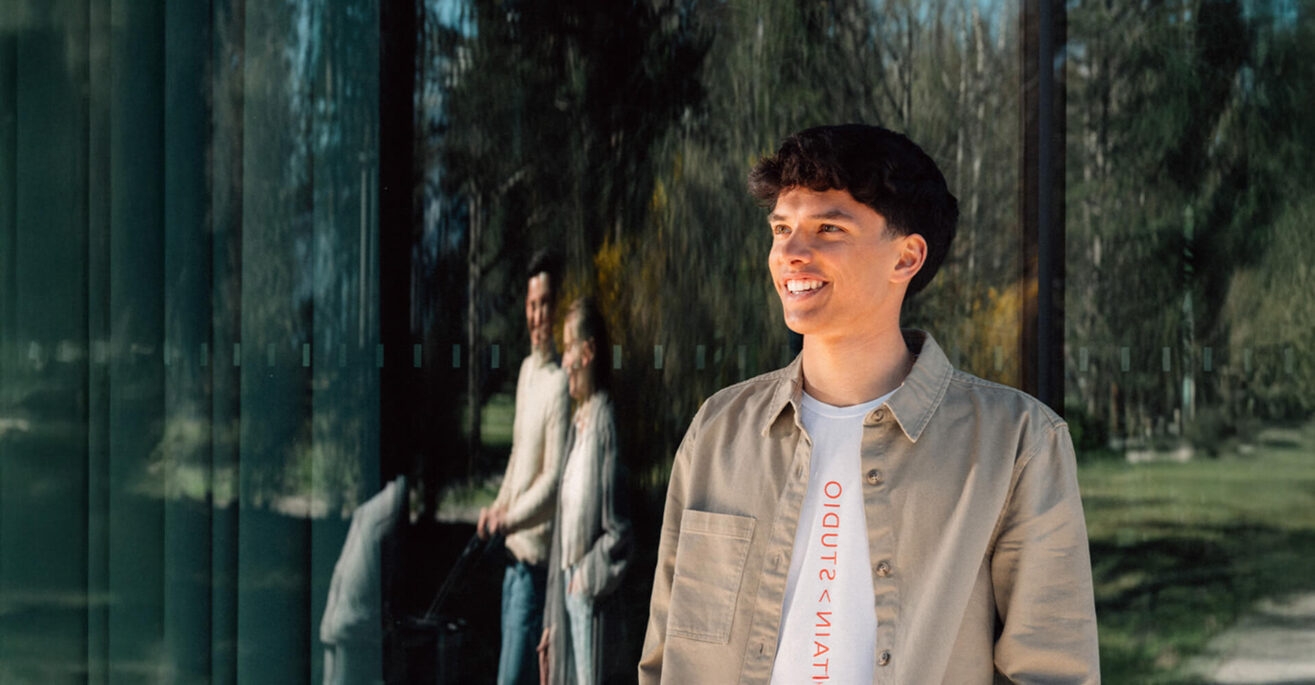 Junger Mann steht lächelnd vor einer Glasfassade, in der das Spiegelbild eines Paares mit Kinderwagen zu sehen ist.