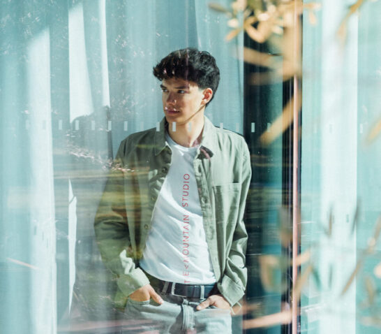 Junger Mann steht hinter einer Glaswand und blickt zur Seite, Lichtreflexionen und Vorhänge im Hintergrund.