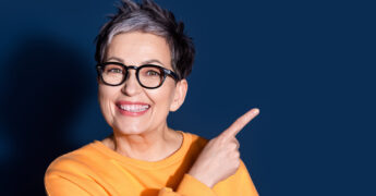 Nahaufnahme einer lachenden Frau mit Brille, die nach rechts zeigt, vor dunkelblauem Hintergrund