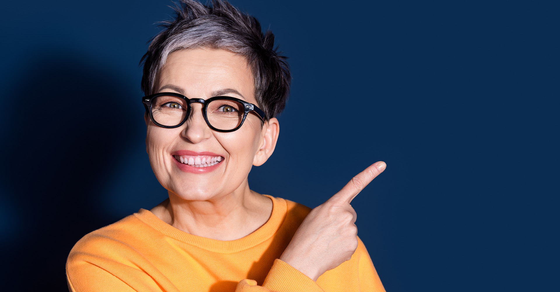 Nahaufnahme einer lachenden Frau mit Brille, die nach rechts zeigt, vor dunkelblauem Hintergrund