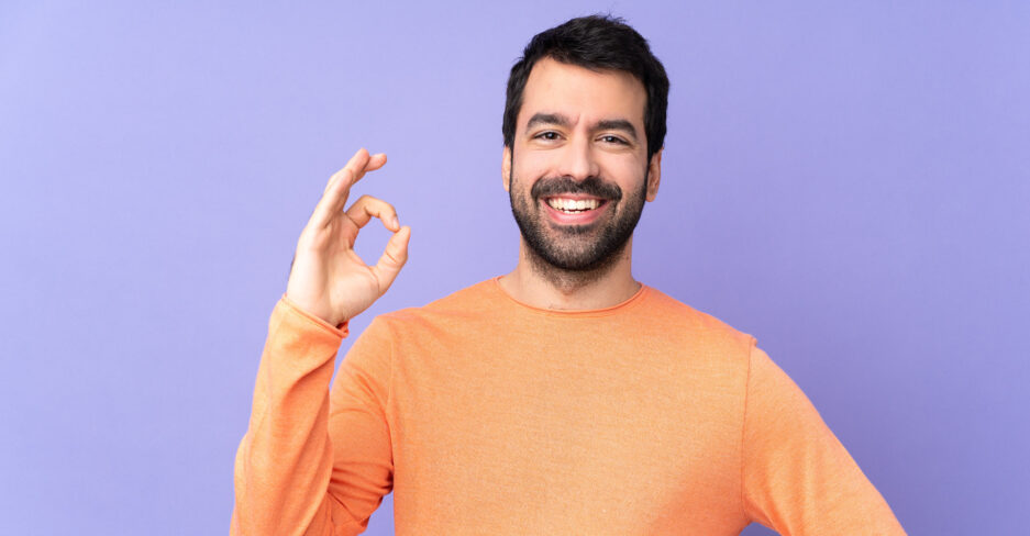 Lächelnder Mann in orangefarbenem Pullover zeigt „Okay“-Geste vor lilafarbenem Hintergrund – Symbol für Zufriedenheit mit dem Gratiskonto