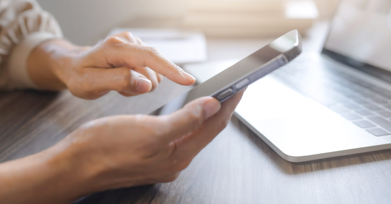 Hand tippt auf Smartphone vor Laptop – ePIN sicher online anzeigen