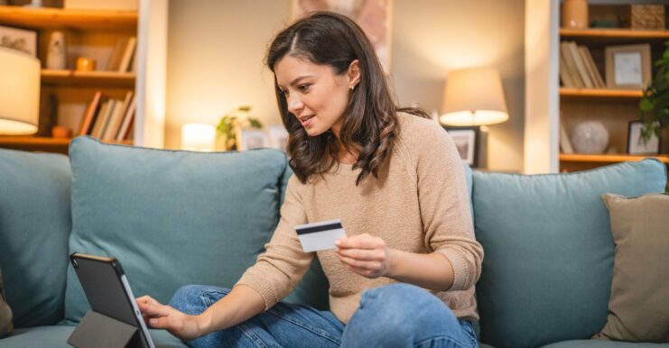 Frau mit Bankomatkarte in der Hand tippt mit rechter Hand auf ein Notebook. Sitzt auf der Couch in einem Wohnzimmer.