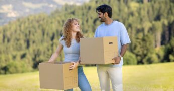 Zwei junge Erwachsene halten einen Umzugkarton in der Hand und stehen auf einem Feld. Im Hintergrund sieht man Bäume und Berge.