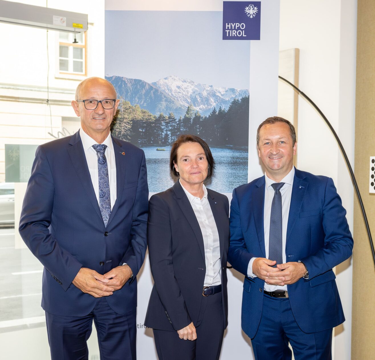 Landeshauptmann Tirol Anton Mattle, Nicole Adrian und Andreas Stadler von der Hypo Tirol stehen im WohnRaum Kompetenzcenter vor einem Rollup mit Hypo Tirol Logo an der rechten oberen Kante und naturaler Kulisse mit See und Berg im Hintergrund.