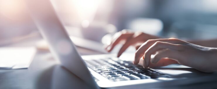 Nahaufnahme von Händen, die auf einer Laptop-Tastatur tippen, im Hintergrund verschwommenes Büro mit hellem Sonnenlicht.