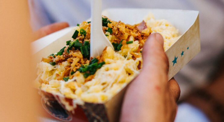 Portion Käsespätzle in Bambusschale mit Röstzwiebeln und Schnittlauch, serviert mit Plastikgabel.