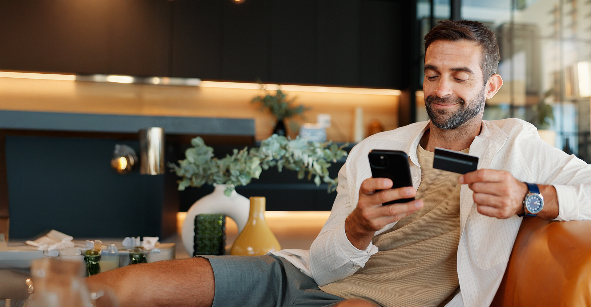 Mann sitzt entspannt auf dem Sofa und bedient sein Smartphone in modernem Wohnzimmer.