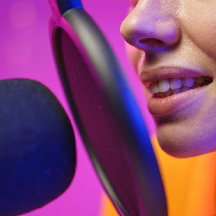 Nahaufnahme eines Mundes vor einem Mikrofon mit Popschutz im Studio, pink-lila Hintergrund.