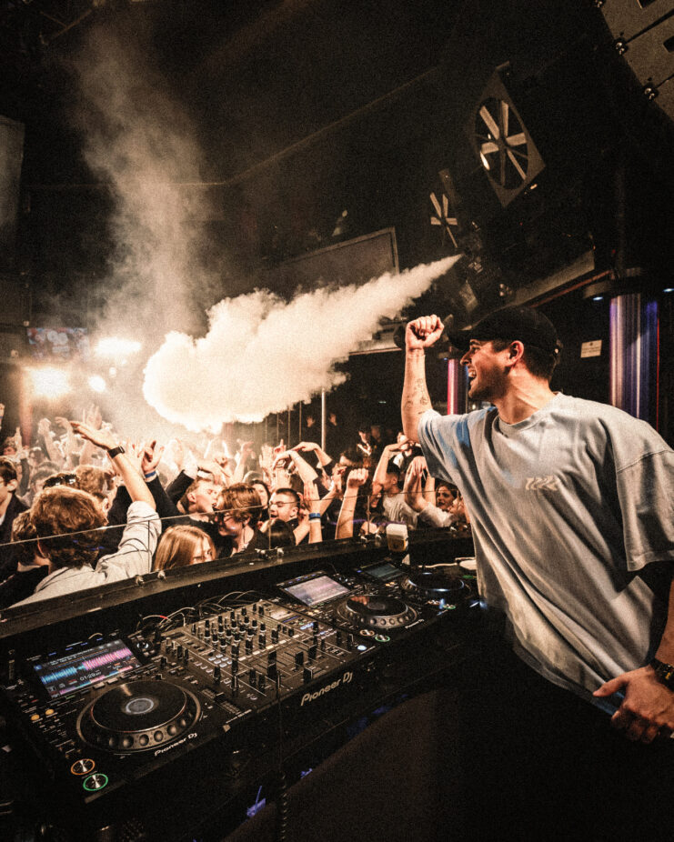 DJ heizt einer feiernden Menge im Club ein, Nebelmaschine und Lichteffekte sorgen für Stimmung.