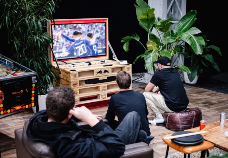 Drei Personen schauen in einer gemütlichen Lounge gemeinsam Fußball auf einem großen Bildschirm.