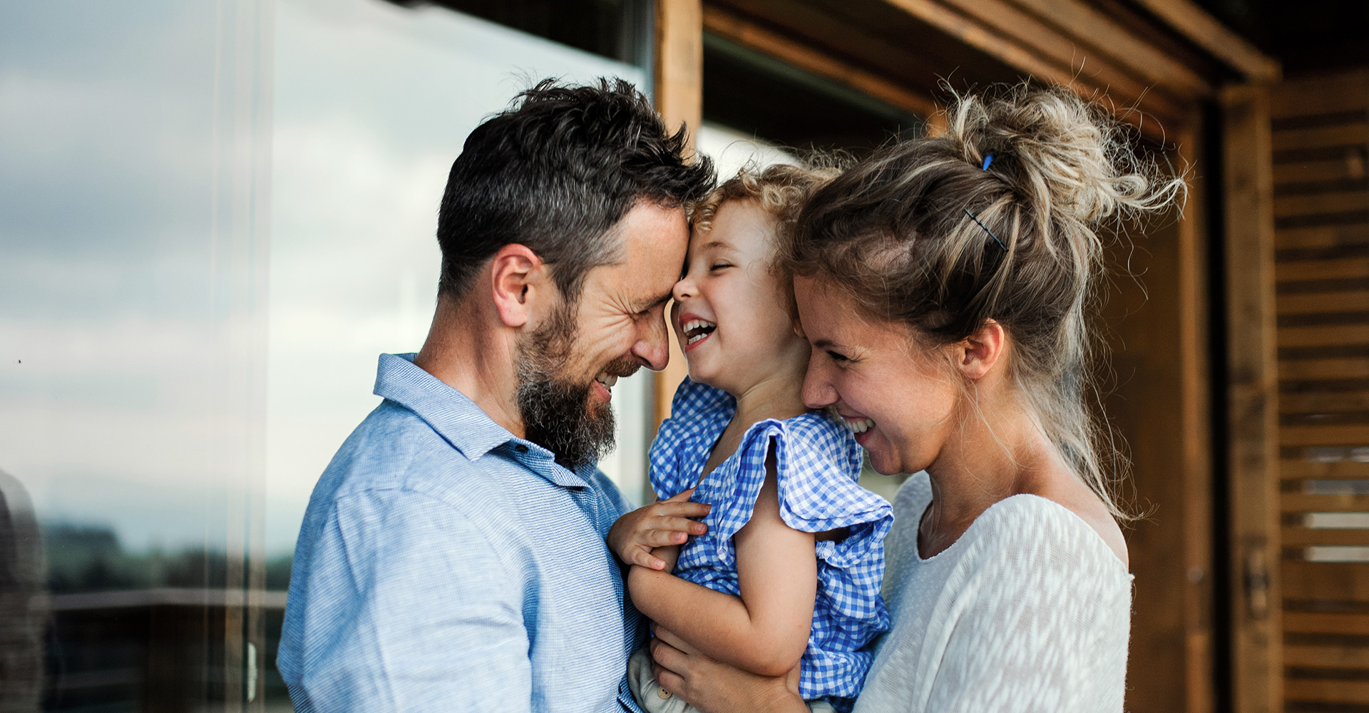 Ein Vater und eine Mutter lachen herzlich mit ihrer kleinen Tochter, die sie im Arm hält. Die Familie steht vor einem Holzhaus und zeigt eine innige, fröhliche Momentaufnahme.