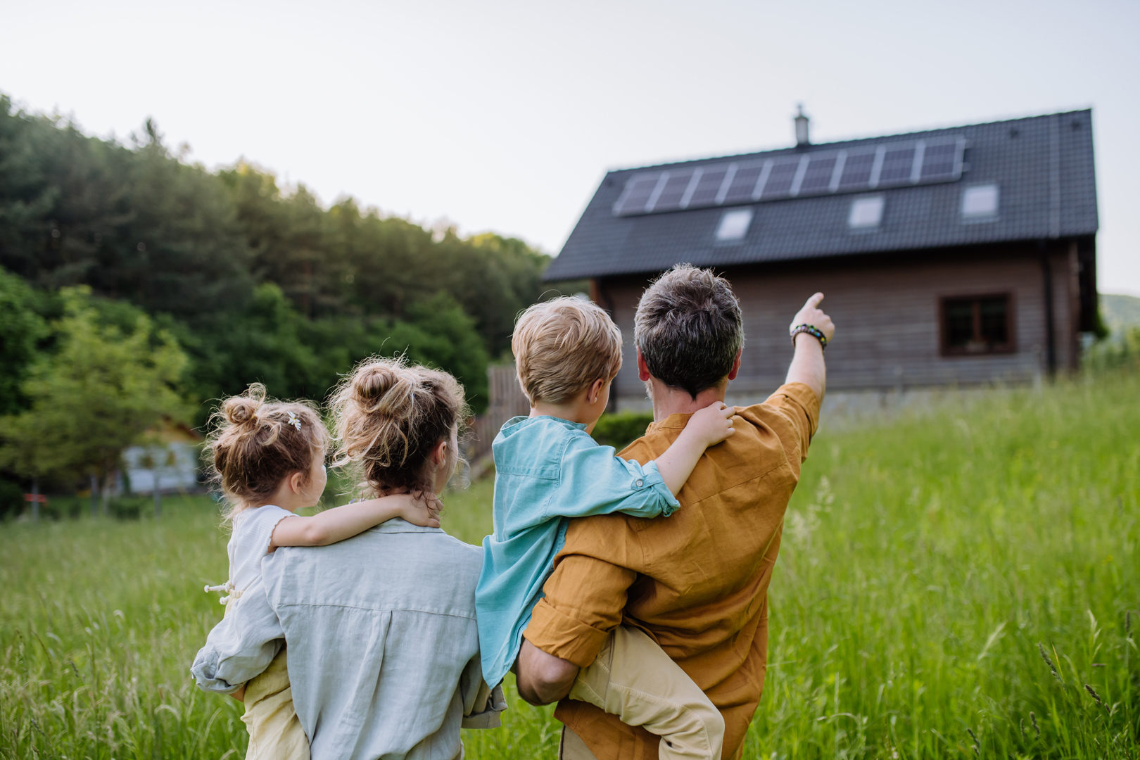 Eine Familie mit zwei Kindern steht auf einer Wiese vor einem Haus mit Solarpaneelen auf dem Dach. Der Vater zeigt nach vorne, während die Kinder und Mutter ihn ansehen. Die Umgebung ist grün und von Bäumen umgeben.
