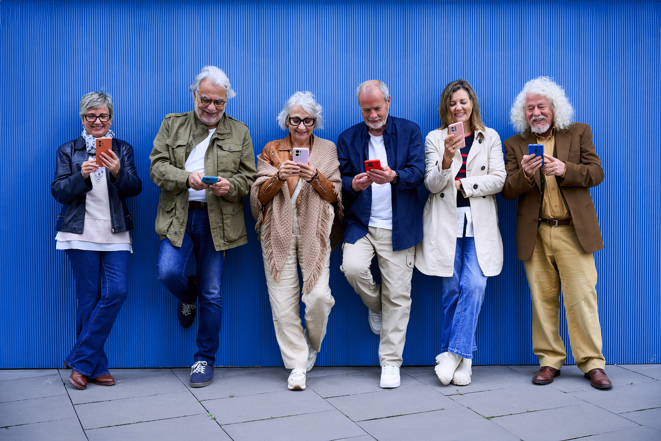Sechs ältere Menschen stehen vor blauer Wand und nutzen ihre Smartphones – Symbol für digitale Teilhabe und moderne Kommunikation