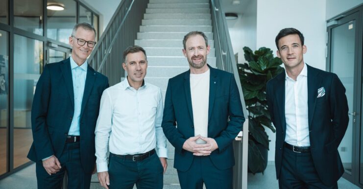 Vier Männer in Businesskleidung stehen vor einer Treppe in einem modernen Büro – Gruppenfoto.