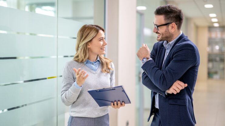 Zwei junge Erwachsene unterhalten sich im Business-Look in einem Bürogebäude. Sie lächeln dabei.