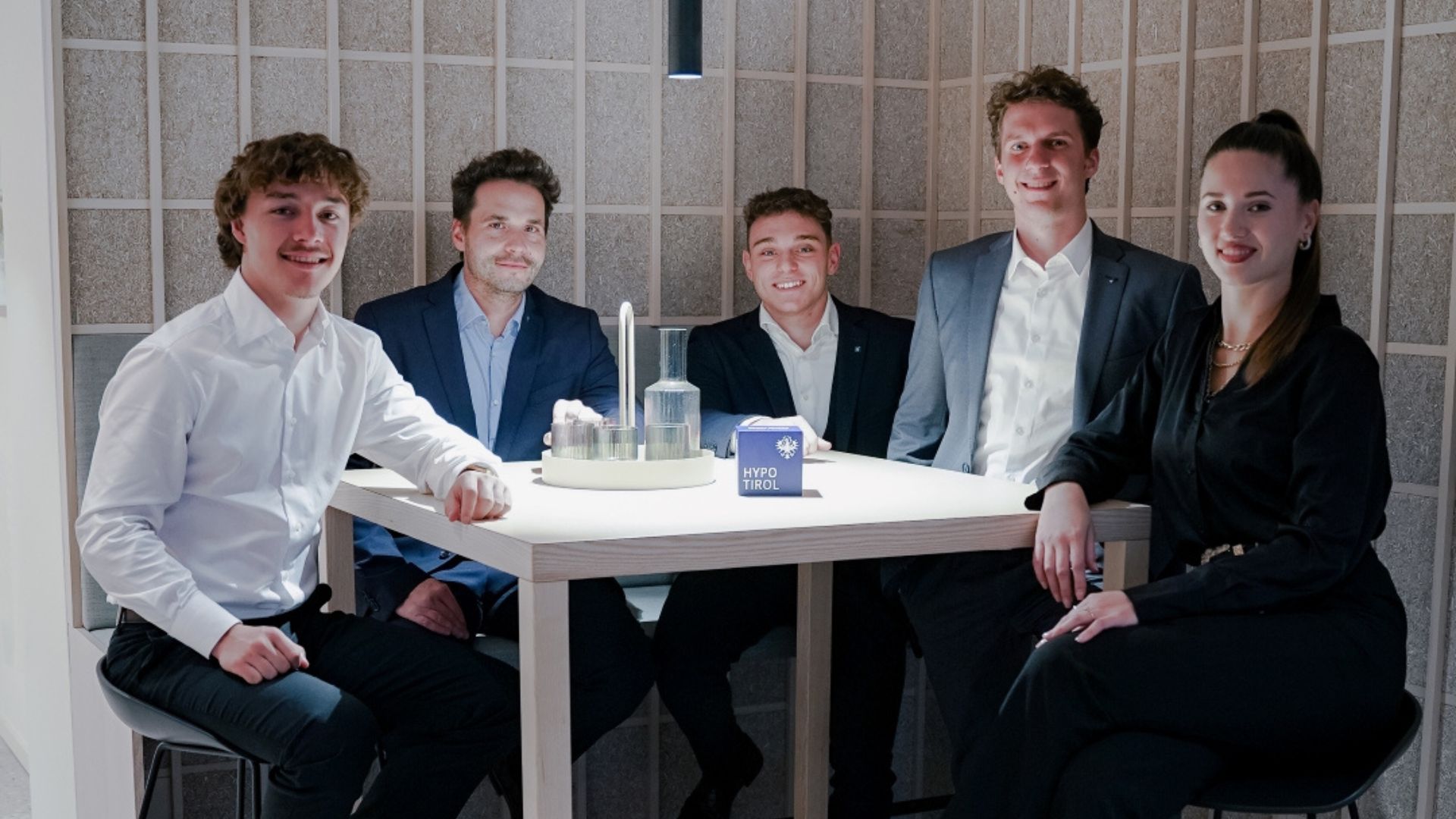 Vier Männer und eine Frau in Businesskleidung posieren lächelnd als junges Team in einem modernen Büro- bzw. Loungebereich.