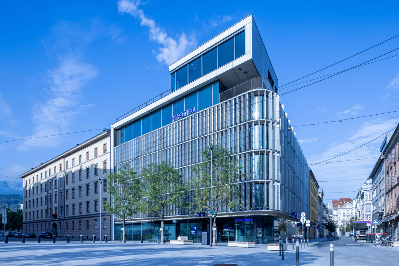 Frontansicht der Hypo Tirol Zentrale in Innsbruck, aufgenommen über den Bozner Platz mit Bäumen im Vordergrund.