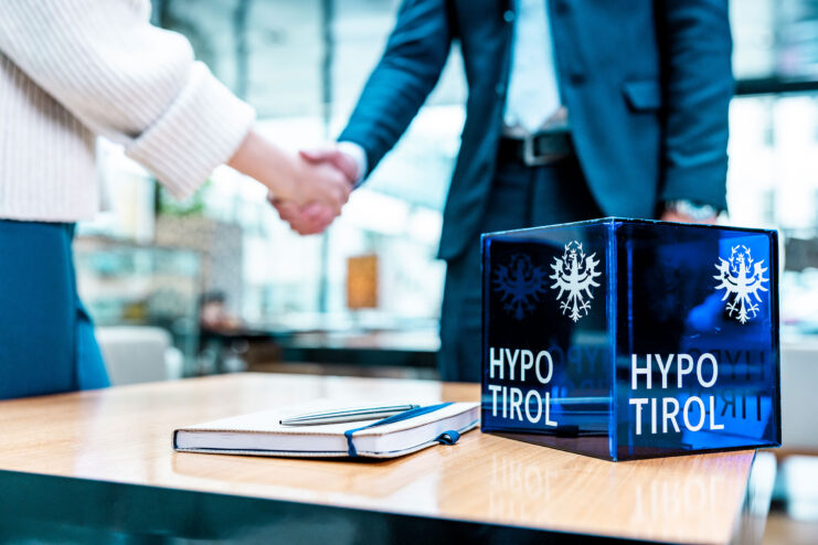 Handschlag im Beratungsgespräch; Hypo Tirol Glaswürfel mit Logo auf dem Tisch, daneben Notizbuch.