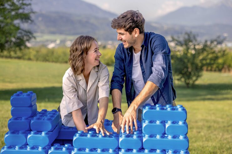 Zwei Personen bauen im Freien gemeinsam eine Konstruktion aus großen blauen Bausteinen, im Hintergrund ist eine Berglandschaft zu sehen.