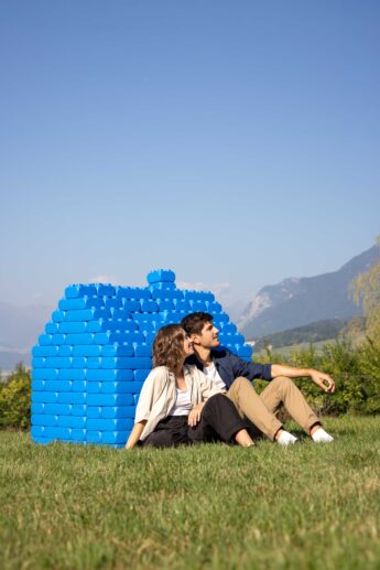Zwei Personen sitzen auf einer Wiese vor einer Mauer aus großen blauen Bausteinen und sehen in die Ferne.