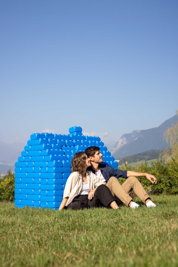 Zwei Personen sitzen auf einer Wiese vor einer Mauer aus großen blauen Bausteinen und sehen in die Ferne.