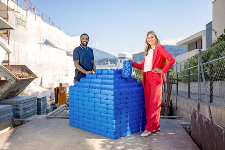 Zwei Personen stehen im Freien neben einer Mauer aus großen blauen Bausteinen.