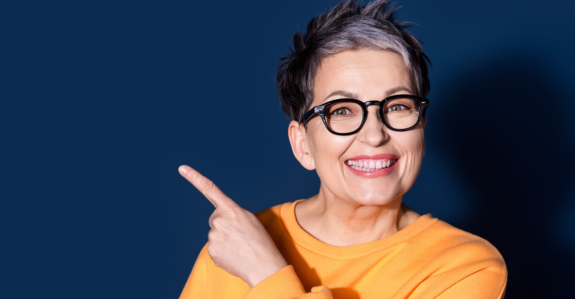 Nahaufnahme einer lachenden Frau mit Brille, die nach rechts zeigt, vor dunkelblauem Hintergrund