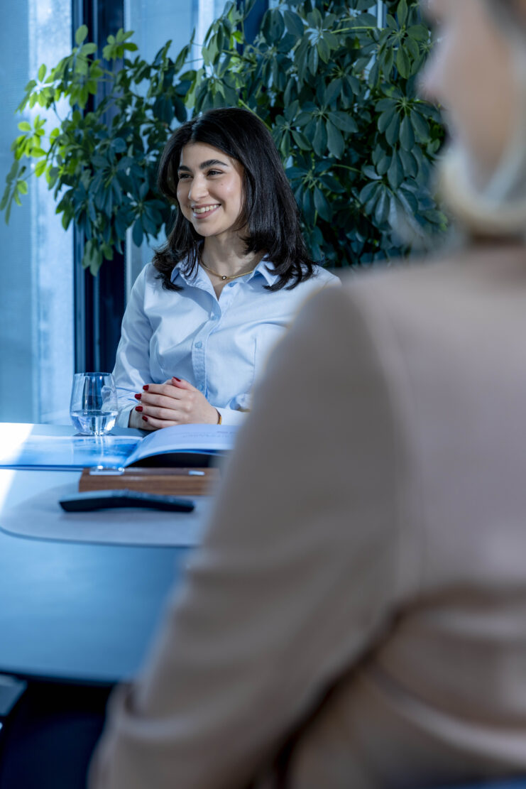 Person sitzt an einem Besprechungstisch in einem Büro und spricht mit einer weiteren Person im Vordergrund.