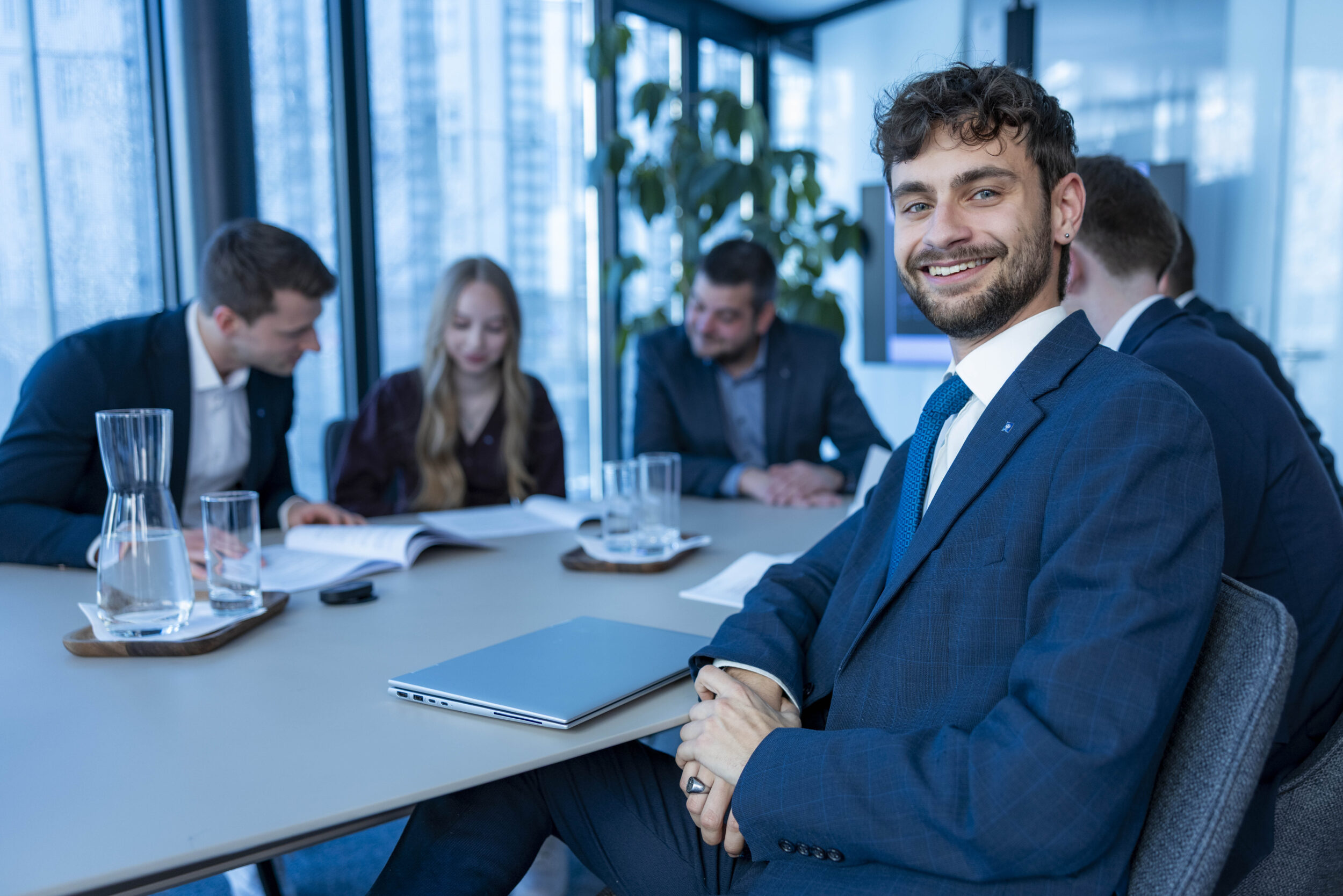 Person sitzt an einem Konferenztisch und blickt seitlich, im Hintergrund arbeiten weitere Personen.