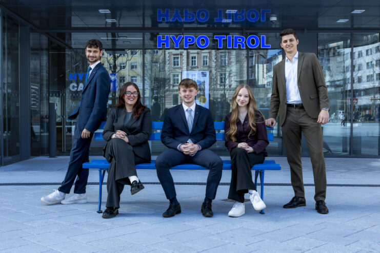 Fünf junge Mitarbeiterinnen und Mitarbeiter der Hypo Tirol stehen und sitzen vor dem Eingang der Hypo Tirol Zentrale in Innsbruck.