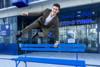 Ein junger Mann in Anzug springt über eine blaue Bank vor der Hypo Tirol.