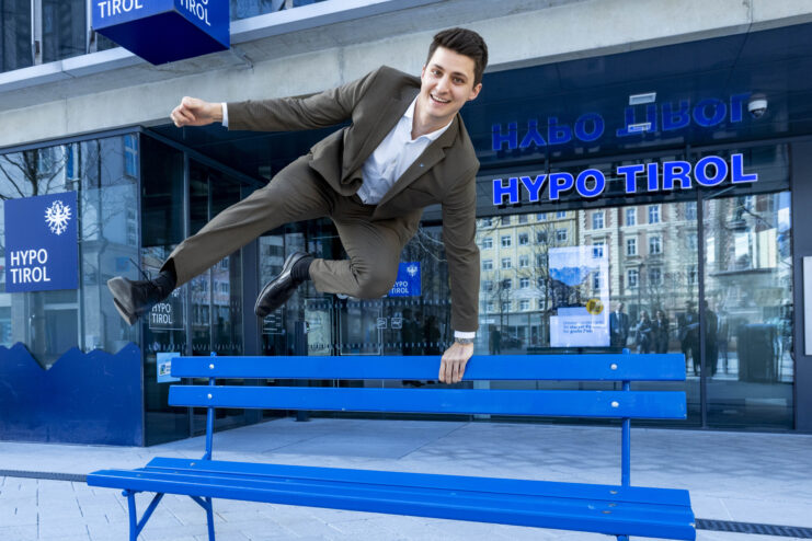 Ein junger Mann in Anzug springt über eine blaue Bank vor der Hypo Tirol.