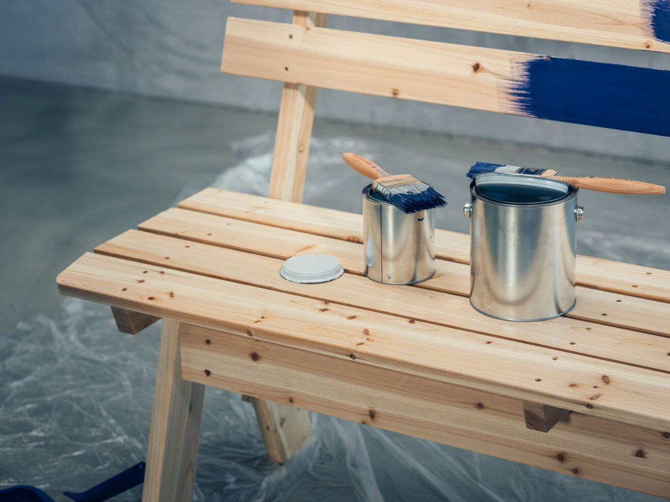 Detailaufnahme einer Holzbank mit Farbeimern, Pinsel und blau gestrichenen Holzteilen.