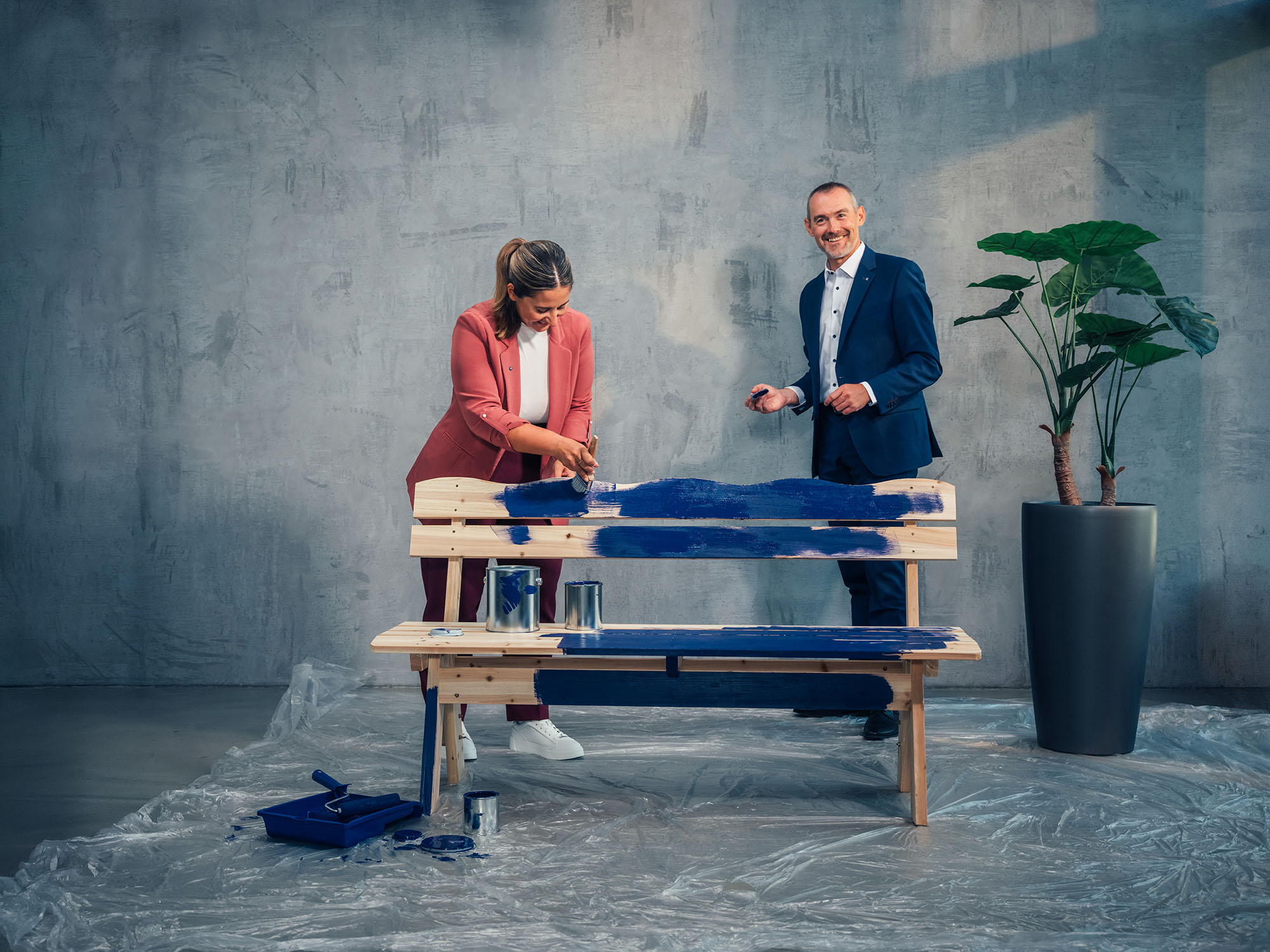 Zwei Personen arbeiten gemeinsam an einer Holzbank und tragen blaue Farbe auf die Holzelemente auf.