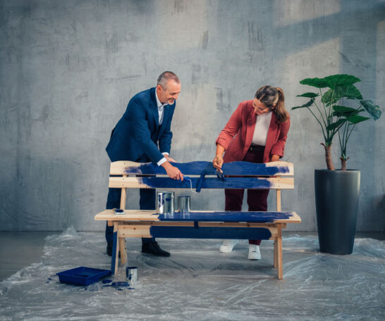 Zwei Personen streichen eine Holzbank mit blauer Farbe, um die Sitzfläche und Rückenlehne zu gestalten.