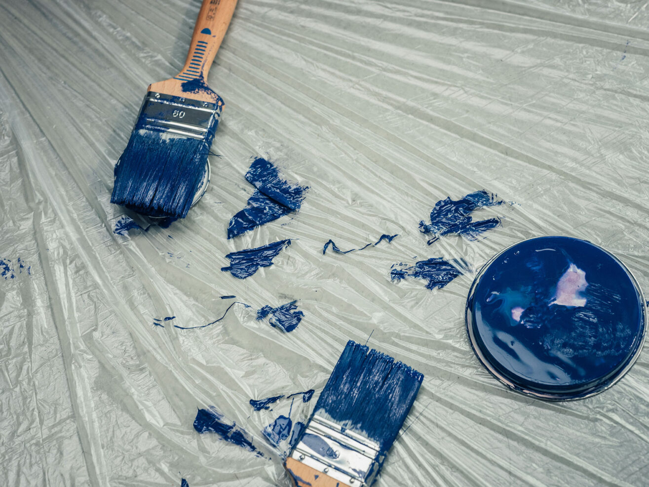 Pinsel, Farbreste und blaue Farbe auf einer mit Folie abgedeckten Arbeitsfläche.