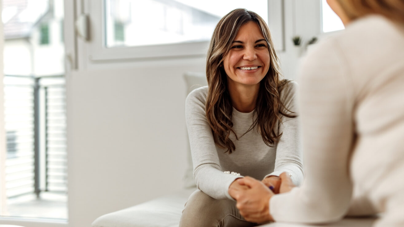Zwei Frauen sitzen einander gegenüber in einem Beratungsraum, halten einander an den Händen und lächeln sich vertrauensvoll an.