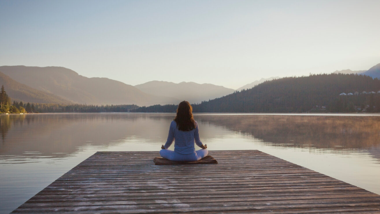 Eine Person sitzt meditierend auf einem Holzsteg an einem ruhigen See, umgeben von Bergen in den Tiroler Alpen, in den frühen Morgenstunden.