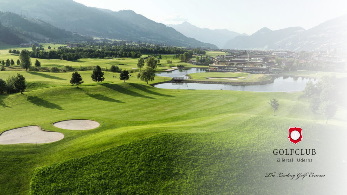 Panoramablick auf einen gepflegten Golfplatz mit Wasserteich, Fairways und Bunkern vor einer alpinen Bergkulisse.