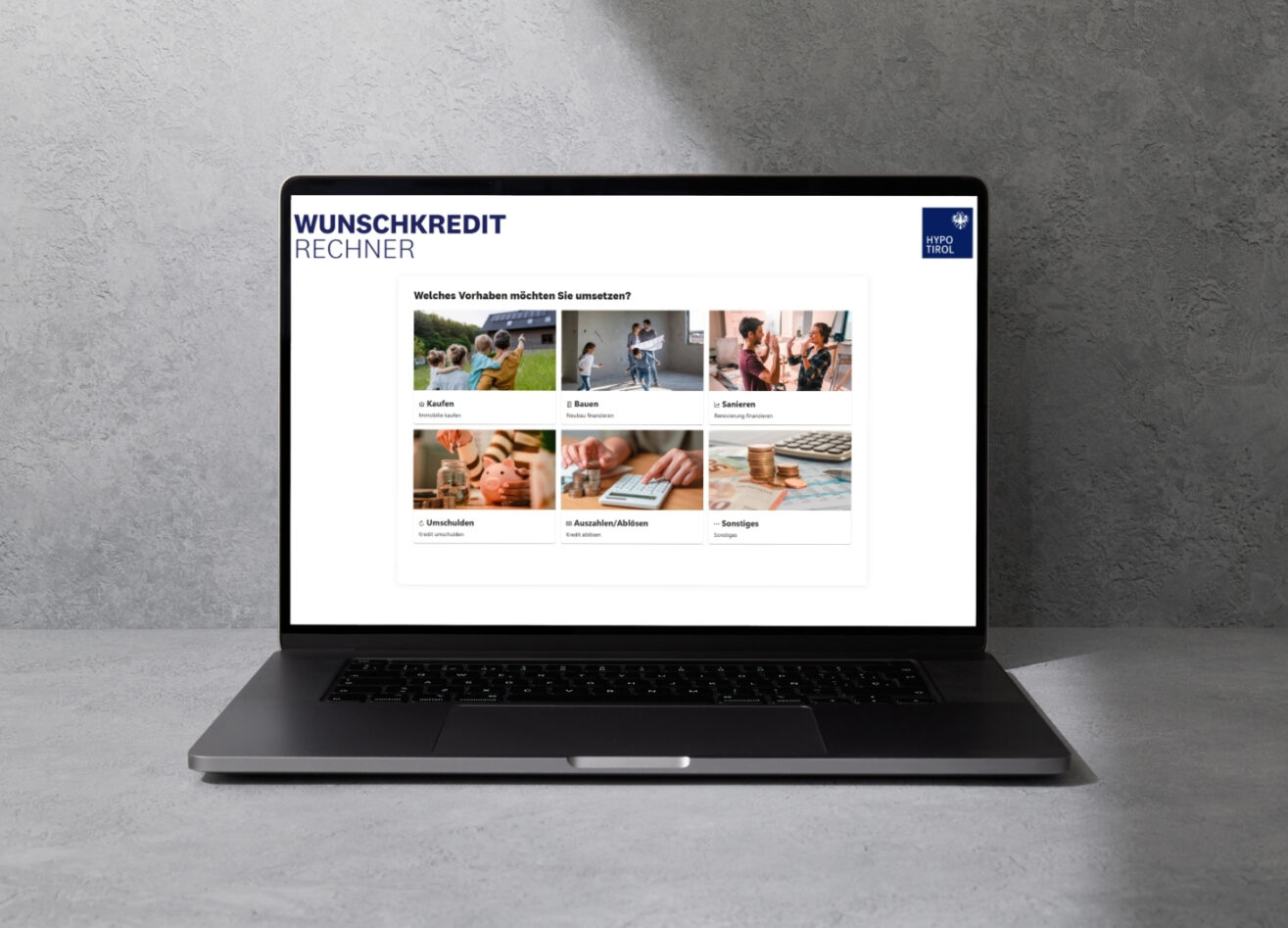 Laptop auf grauer Oberfläche, auf dessen Bildschirm ein Wunschkredit‑Rechner mit verschiedenen Auswahlfeldern für Finanzierungszwecke angezeigt wird.