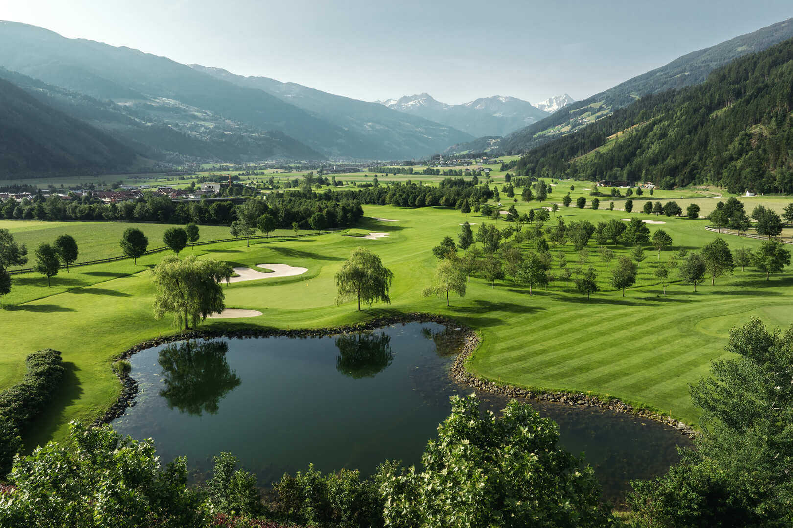 Panoramablick auf einen gepflegten Golfplatz mit Wasserteich, Fairways und Bunkern vor einer alpinen Bergkulisse.