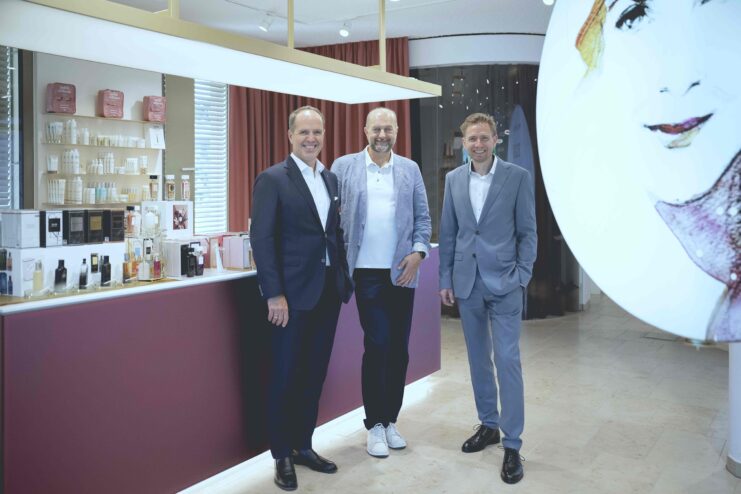 Drei Personen stehen in einem modernen Geschäfts- bzw. Showroom-Bereich mit Kosmetik- und Pflegeprodukten. Im Hintergrund befinden sich Regale mit verschiedenen Produkten, rechts ist ein großformatiges Porträtmotiv zu sehen. Die Aufnahme zeigt einen Unternehmensbesuch.