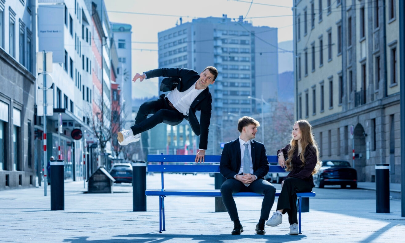 Drei junge Mitarbeiterinnen und Mitarbeiter der Hypo Tirol auf einer blauen Bank in Innsbruck; eine Person springt dynamisch über die Bank, während zwei weitere sitzend miteinander sprechen.