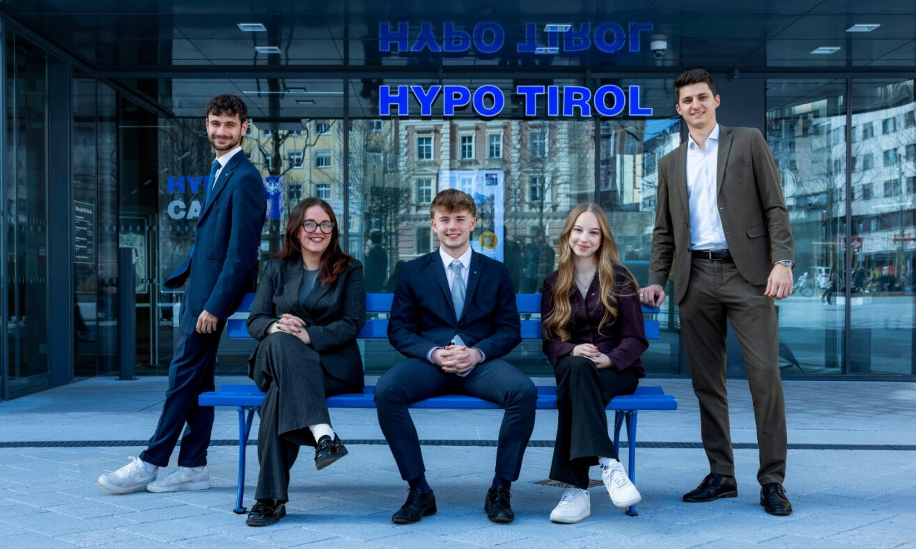 Fünf junge Mitarbeiterinnen und Mitarbeiter der Hypo Tirol stehen und sitzen vor dem Eingang der Hypo Tirol Zentrale in Innsbruck.