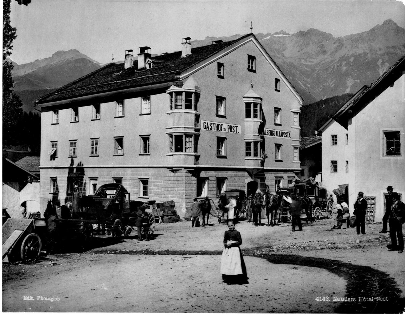 Schwarz-Weiß-Fotografie des Gasthof zur Post in Nauders. Ein Mädchen in einem Kleid mit weißer Schürze im Vordergrund, Pferdegespanne vor dem Hotel.