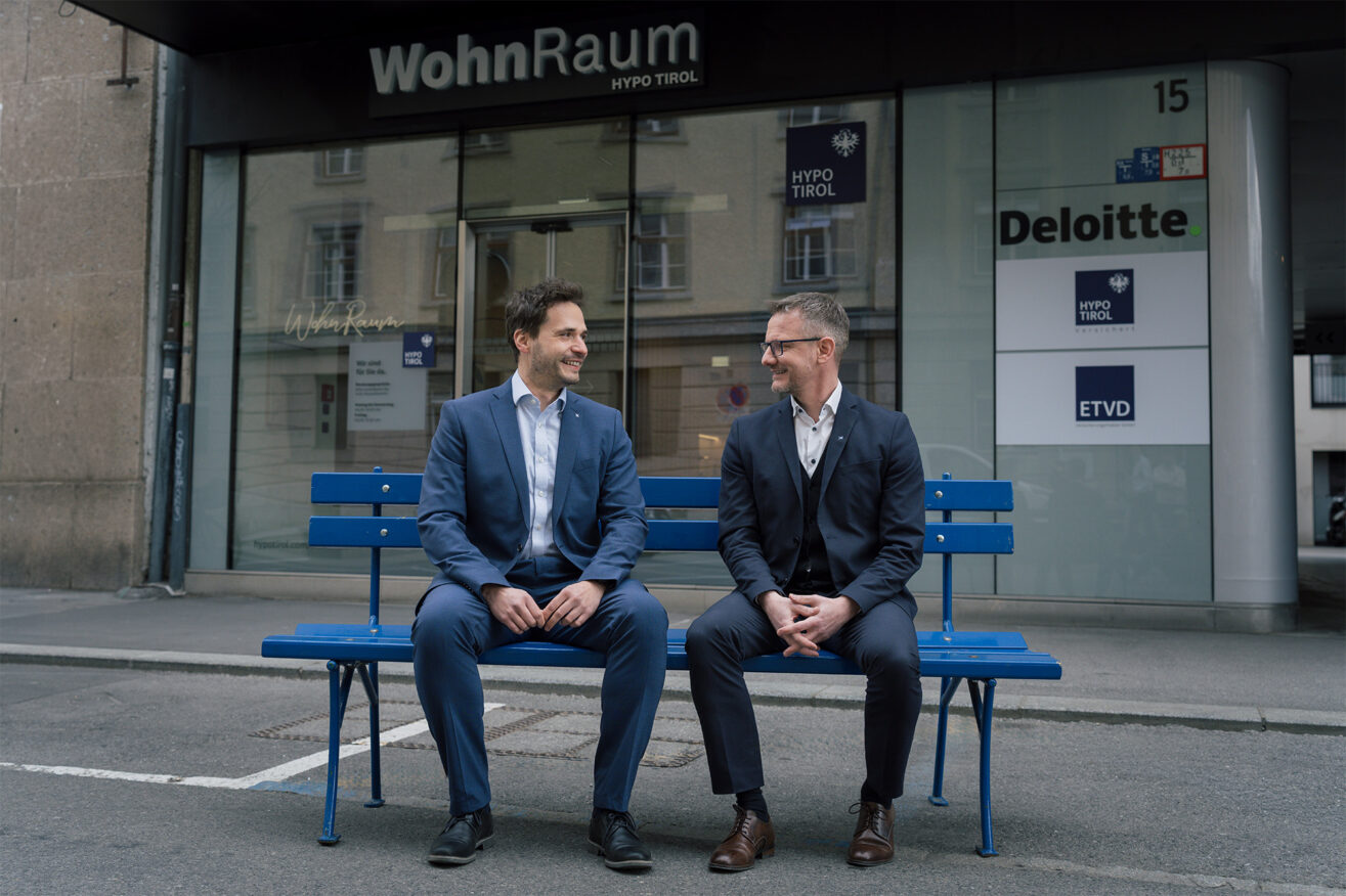 Zwei WohnRaum-Berater der Hypo Tirol auf der hypoblauen Bank vor dem WohnRaum-Kompetenzcenter